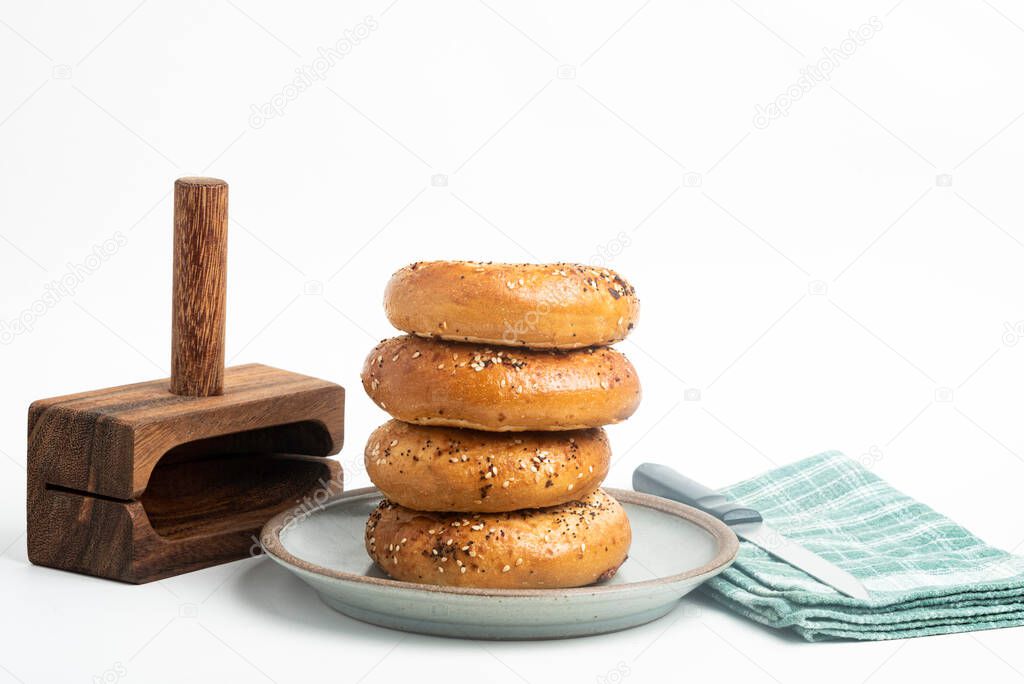 A single tall stack of four freshly baked bagels on a ceramic plate with a cutting wood stand and knife set on a plain white background.