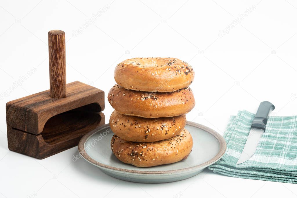 A single tall stack of four freshly baked bagels on a ceramic plate with a cutting wood stand and knife set on a plain white background.