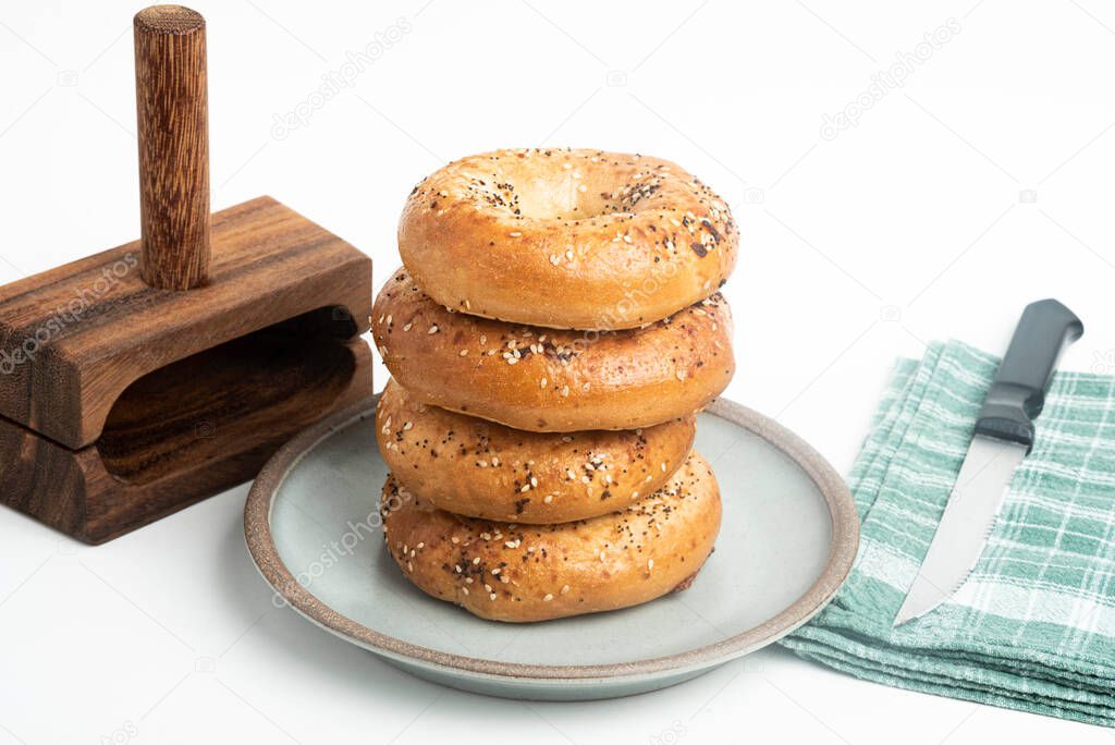 A single tall stack of four freshly baked bagels on a ceramic plate with a cutting wood stand and knife set on a plain white background.