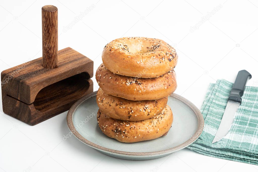 A single tall stack of four freshly baked bagels on a ceramic plate with a cutting wood stand and knife set on a plain white background.