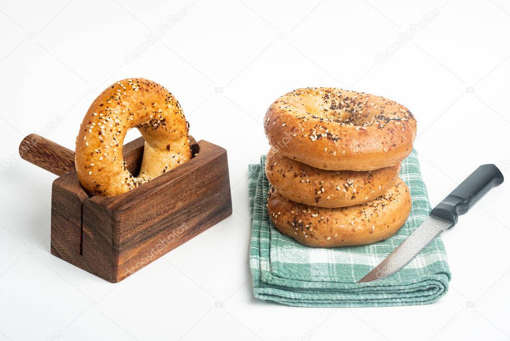 A single tall stack of three freshly baked bagels on a napkin with one bagel on a cutting wood stand and knife set on a plain white background.