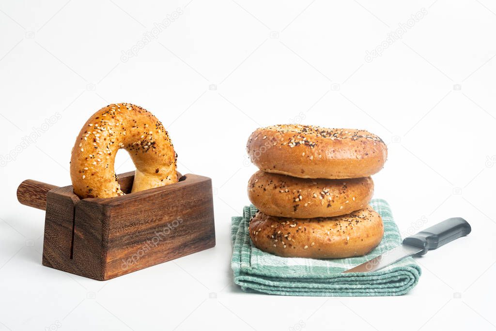 A single tall stack of three freshly baked bagels on a napkin with one bagel on a cutting wood stand and knife set on a plain white background.