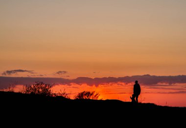 Kırmızı gökyüzünün arka planında insanların siluetleri. Deniz kıyısında gün batımı, insanlar bir tepede yürüyorlar. Akşam. Bahar.