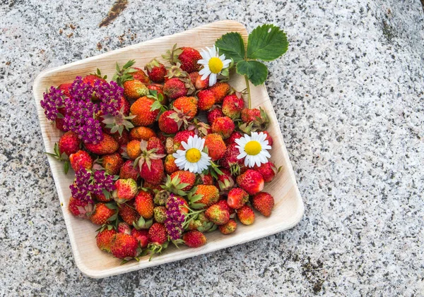 Tahta bir tabakta taze kırmızı çilek. Çilek C vitamini kaynağıdır. Bu meyve doğal bir afrodizyak. Doğal granit taş arka planda kır çiçekleri ile kolaj.