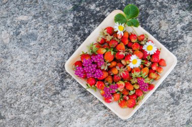 Tahta bir tabakta taze kırmızı çilek. Çilek C vitamini kaynağıdır. Bu meyve doğal bir afrodizyak. Doğal granit taş arka planda kır çiçekleri ile kolaj.