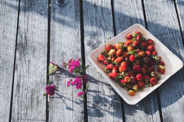 Tahta bir tabakta taze kırmızı çilek. Çilek C vitamini kaynağıdır. Bu bir berry - doğal bir afrodizyak, eski ahşap tahtalardan masanın arka planda yabani çiçekler ile Kolaj.