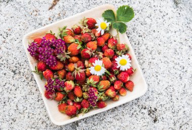 Tahta bir tabakta taze kırmızı çilek. Çilek C vitamini kaynağıdır. Bu meyve doğal bir afrodizyak. Doğal granit taş arka planda kır çiçekleri ile kolaj.