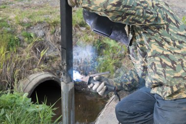 Kaynakçı işte. Kaynak makinesi kullanılarak metal bir kolon montajı. Elektrotlardan kıvılcımlar. Yaz. Gün.