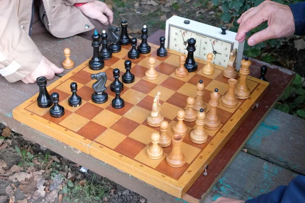 Two men play chess. Wooden chess pieces on a wooden board. Beginning of the party, debut. The game takes place against the clock. Park bench. Autumn day.