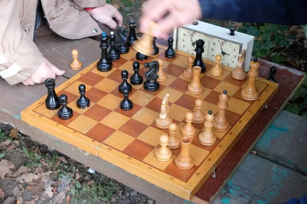 Two men play chess. Wooden chess pieces on a wooden board. Beginning of the party, debut. The game takes place against the clock. Park bench. Autumn day.