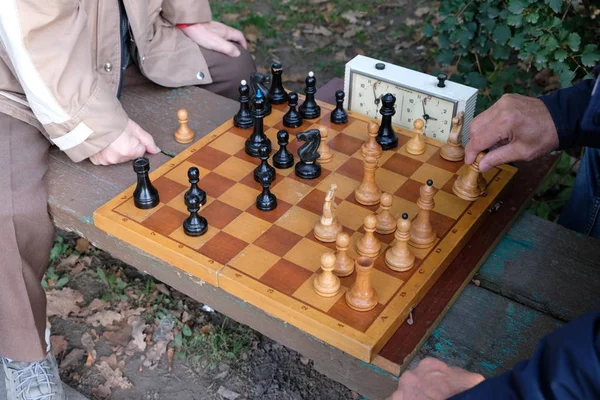 Two men play chess. Wooden chess pieces on a wooden board. Beginning of the party, debut. The game takes place against the clock. Park bench. Autumn day.