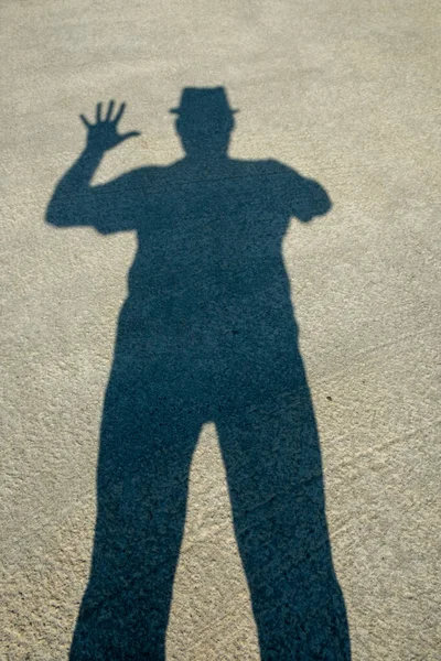 Shadow of a man in a hat on the concrete floor. Human hands show symbols Dark silhouette from shadow. Signs: Okay, five, there, victory. Day. Sunny. Georgia.