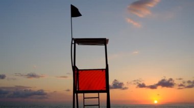 Deniz kenarındaki kurtarma kulesi. Suyun yanındaki kırmızı gözlem noktası. İşaret, kırmızı bayrak. Rüzgar kırmızı bayrağı dalgalandırıyor. Sancak rüzgardan titriyor. Georgia mı? İyi günler, güneşli. Yaz.