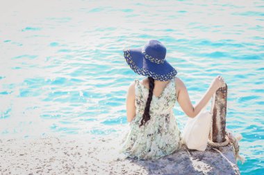 Denize yakın iskelede tek başına oturan hasır şapkalı kadının arka görünümü.