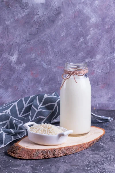 Vegan rice milk in a glass bottle and rice in a white plate on a wooden stand on a gray background with a gray napkin