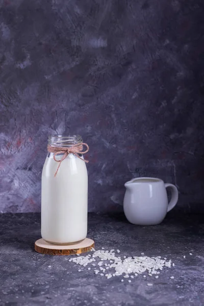Vegan rice milk in a glass bottle and rice in a white plate on a wooden stand with a molon on a gray background