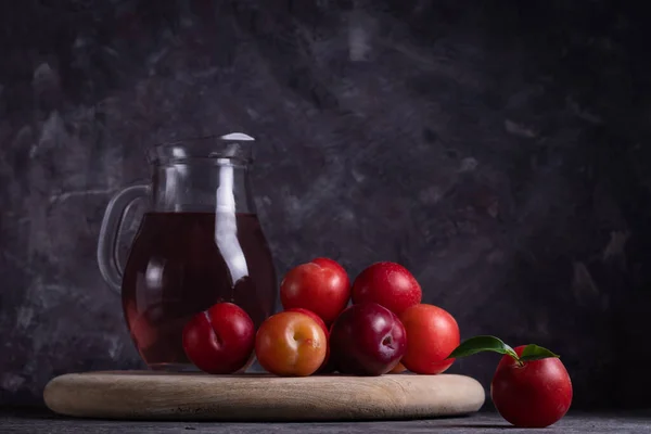 Juice or stewed fruit of ripe red plum in a glass jug on a wooden stand on a gray background in low key