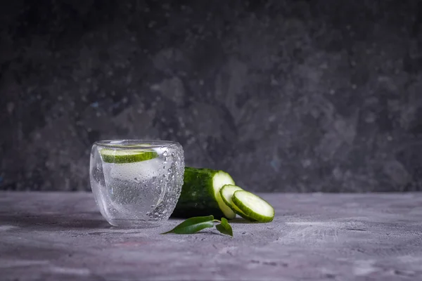 Refreshing water with ice and cucumber in a glass cup with dew on a gray background