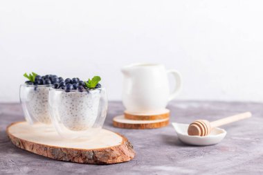 Naneli Chia pudingi ve gözlüklü yaban mersini, ahşap bir tezgahta bal çubuğu ve krema sürahisi ile ışıklı arka planda.