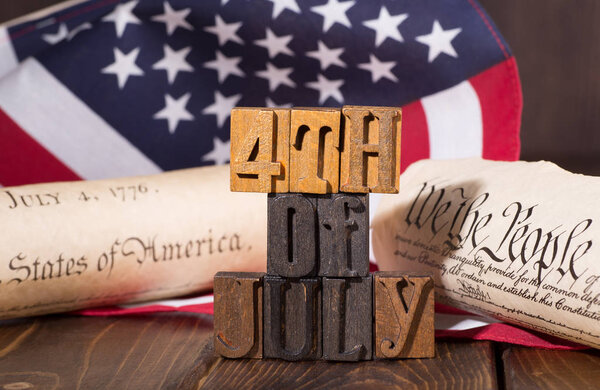"Знак "4 июля" с использованием деревянных буквенных блоков с флагом США, провозглашением независимости и конституцией в фоновой американской праздничной концепции
