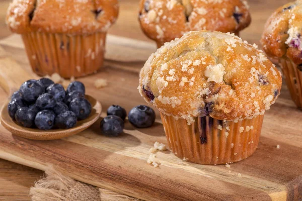 Closeup of a blueberry muffin and spoonful of berries on a wooden board with muffins in background