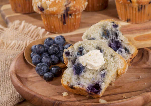 Sliced blueberry muffin and spoonful of berries on a wooden plate with muffins in background