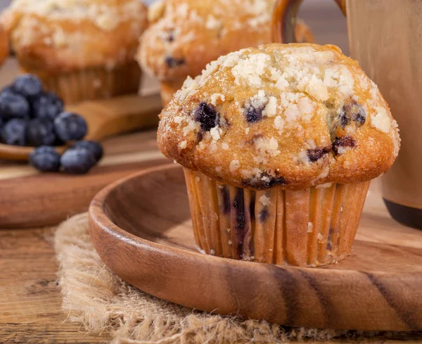 Closeup of a blueberry muffin on a wooden plate with blueberries and muffins in background