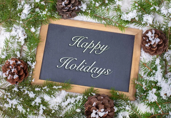 Chalkboard with Happy Holiday text bordered with snow covered pine cones and evergreen tree branches