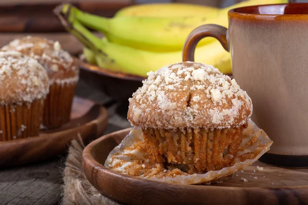 Closeup of a banana nut muffin with a coffee cup on a wooden plate with muffins and bananas in background