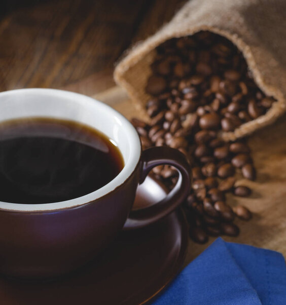 Closeup of a steaming cup of coffee with coffee beans spilled from a burlap bag in background