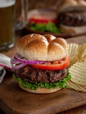 Marullu, domatesli ve soğanlı sulu ızgara hamberger ekmeğin üstünde patates cipsi ve ahşap tahta üzerinde patates cipsi.