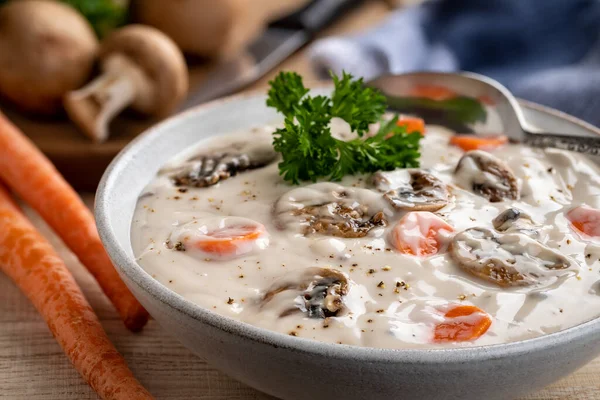 Closeup of bowl of creamy mushroom soup and carrots with ingredients on rustic wooden table.