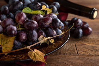 Bir tabakta mor üzümler ve kırsal bir masada arka planda şarap şişesi.