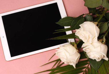 Beyaz bir yüzey üzerinde yatan beyaz güller ve yapraklar. boş ekranlı tablet. Soluk şeftali arka planda üst görünümü kadar stilde mock.