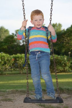 Sırt çantası dik duran ve kameraya bakarak sallanan 5 yaşındaki kızıl saçlı mutlu çocuk