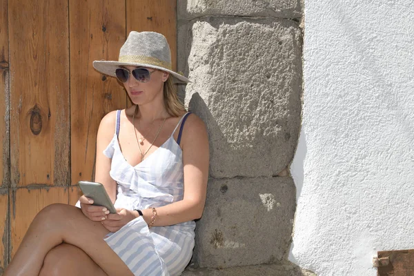 Woman sits under the sun with sunglasses and a hat using her smart phone. With a wooden door and a stoned and white background — Stock Photo, Image