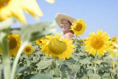 Şapkası olan orta yaşlı bir kadın gülümsüyor ve ayçiçeği tarlasındaki çiçeklerin arasında kameraya bakıyor. Aşağıdan görüntüle