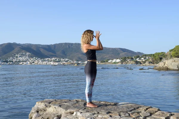 Young girl doing yoga on a small beach pier with a Mediterranean coastal town in the background. Staying fit. Pranamasana yoga posture — Stock Photo, Image