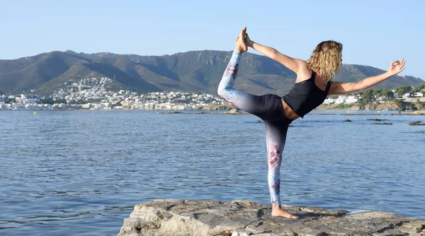 Young girl doing yoga on a small beach pier with a Mediterranean coastal town in the background. Staying fit. With copy space. Natarajasana yoga posture — Stock Photo, Image