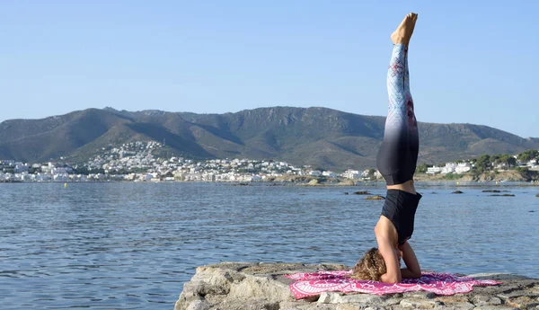 Genç kız arka planda bir Akdeniz sahil kasabası ile küçük bir plaj iskelesinde yoga yapıyor. Formda kalmak. Kopyalama alanı ile. Salamba Sirsasana yoga duruşu