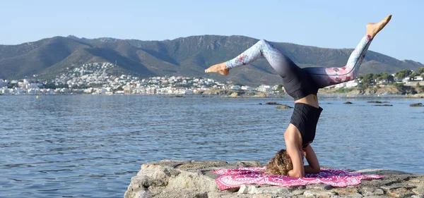 Genç kız arka planda bir Akdeniz sahil kasabası ile küçük bir plaj iskelesinde yoga yapıyor. Formda kalmak. Kopyalama alanı ile. Salamba Sirsasana yoga duruş varyasyonu