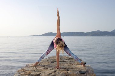 Genç kız arka planda bir Akdeniz sahil kasabası ile küçük bir plaj iskelesinde yoga yapıyor. Formda kalmak