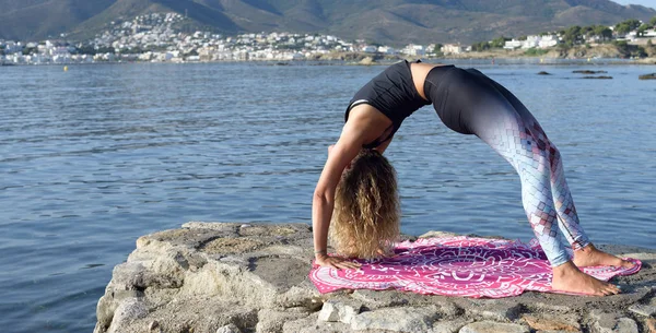 Genç kız arka planda bir Akdeniz sahil kasabası ile küçük bir plaj iskelesinde yoga yapıyor. Formda kalmak. Kopyalama alanı ile. Urdhva Dhanurasana yoga duruşu — Stok fotoğraf