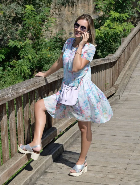 Mulher loira usando óculos de sol e vestido impresso de cor clara em uma ponte de madeira falando ao telefone e sorrindo em um ambiente natural — Fotografia de Stock