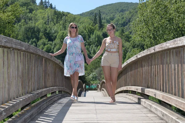 Two women holding hands walk on a wooden bridge in a natural environment