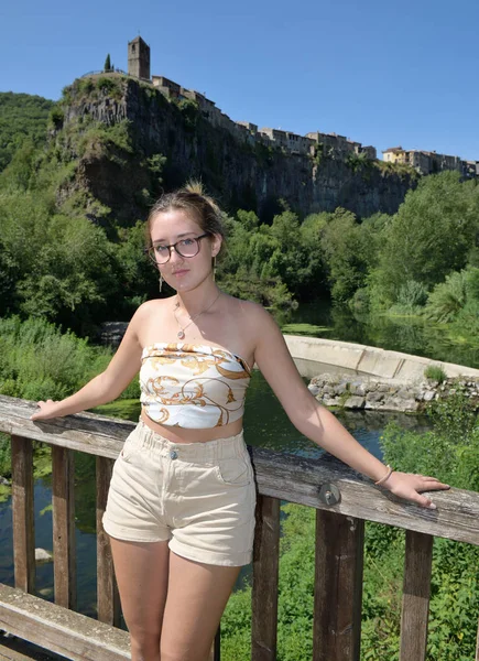 Blondýnka na dřevěném mostě, který hledí na kameru a podpíná zábradlí v přírodním prostředí se středověkou vesnicí v pozadí — Stock fotografie