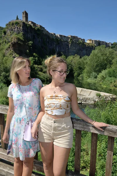 Dvě ženy na dřevěném mostě podporovaly zábradlí v přírodním prostředí se středověkou obcí na pozadí zvané Castellfollit de La Roca v Katalánsku — Stock fotografie