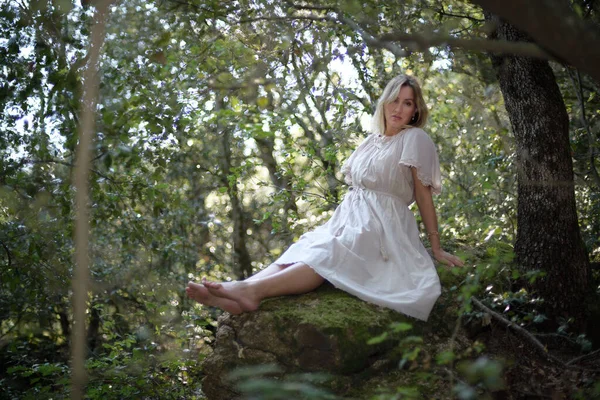 Retrato de una mujer descalza con un vestido rústico sentada sobre una piedra en un bosque oscuro mirando a la cámara — Foto de Stock