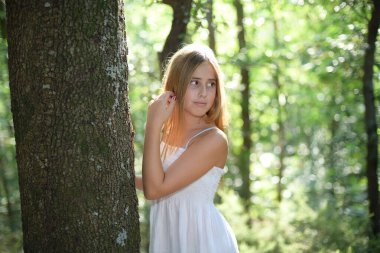 Ormandaki bir ağaca yaslanan beyaz elbiseli bir kız.