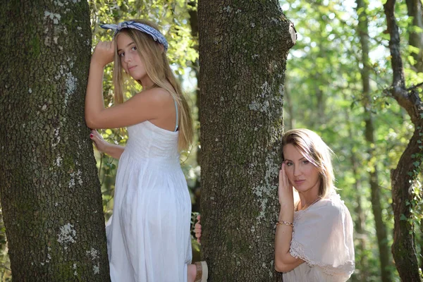 Madre e hija apoyadas en un árbol en un bosque con vestidos claros —  Fotos de Stock